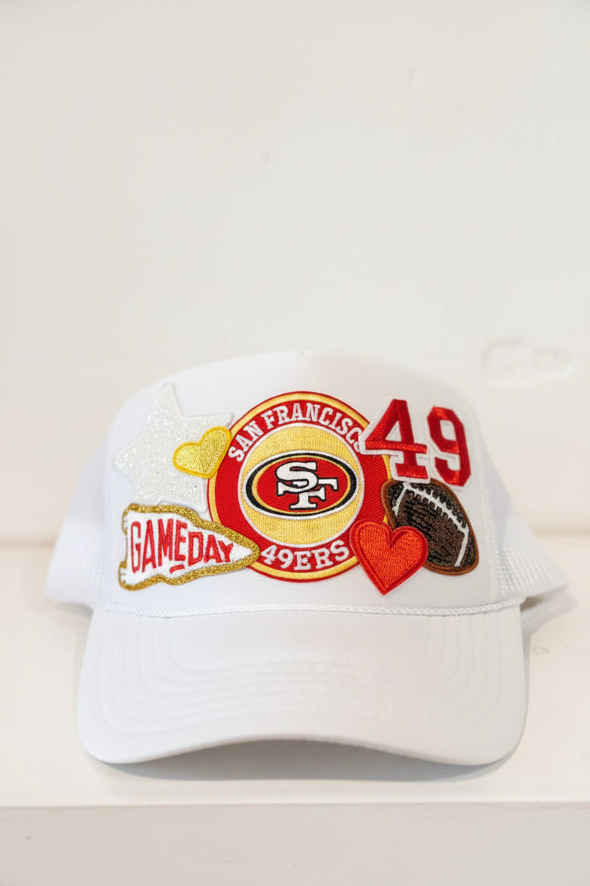 A white San Francisco 49ers trucker hat with embroidered patches on the front, including the team logo, the number 49, a red heart, a football patch, and a glittery star patch, set against a plain off-white background.