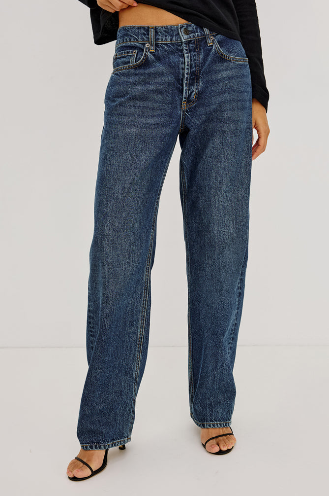 A person wearing blue straight-leg denim jeans with a black long-sleeve top and black heeled sandals, shown from the waist down against a plain light gray studio background.